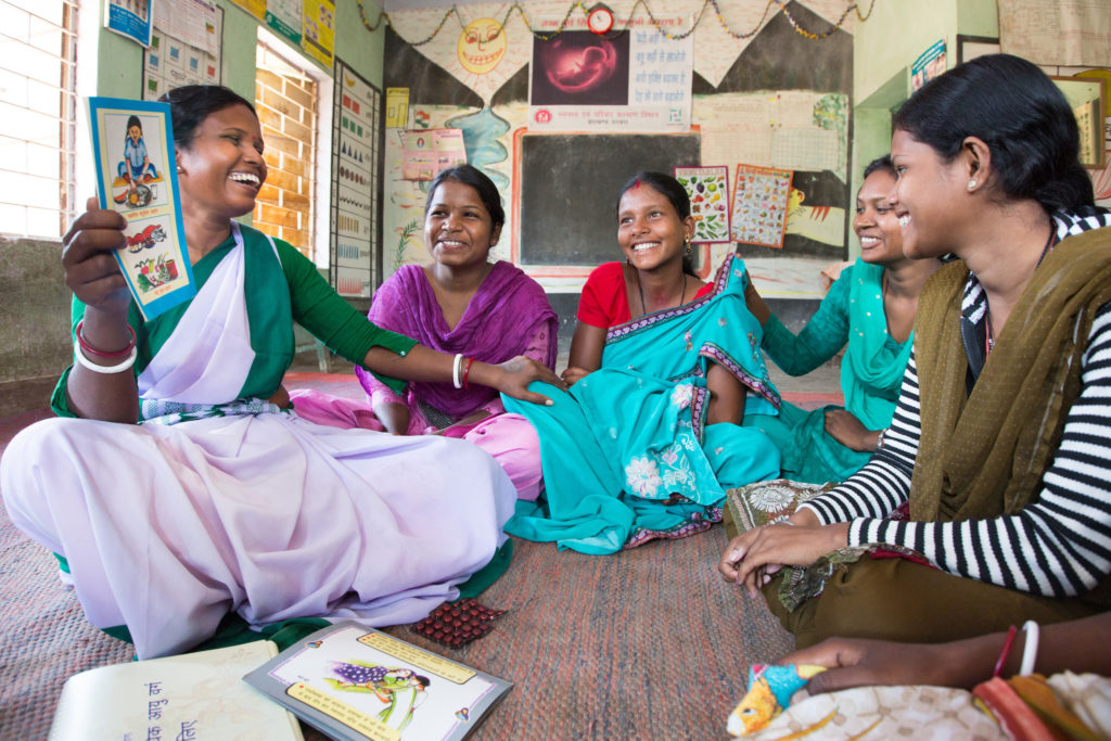 Five smiling women sit on the floor of a classroom in a semi-circle. The woman on the left holds up a diagram from a book.