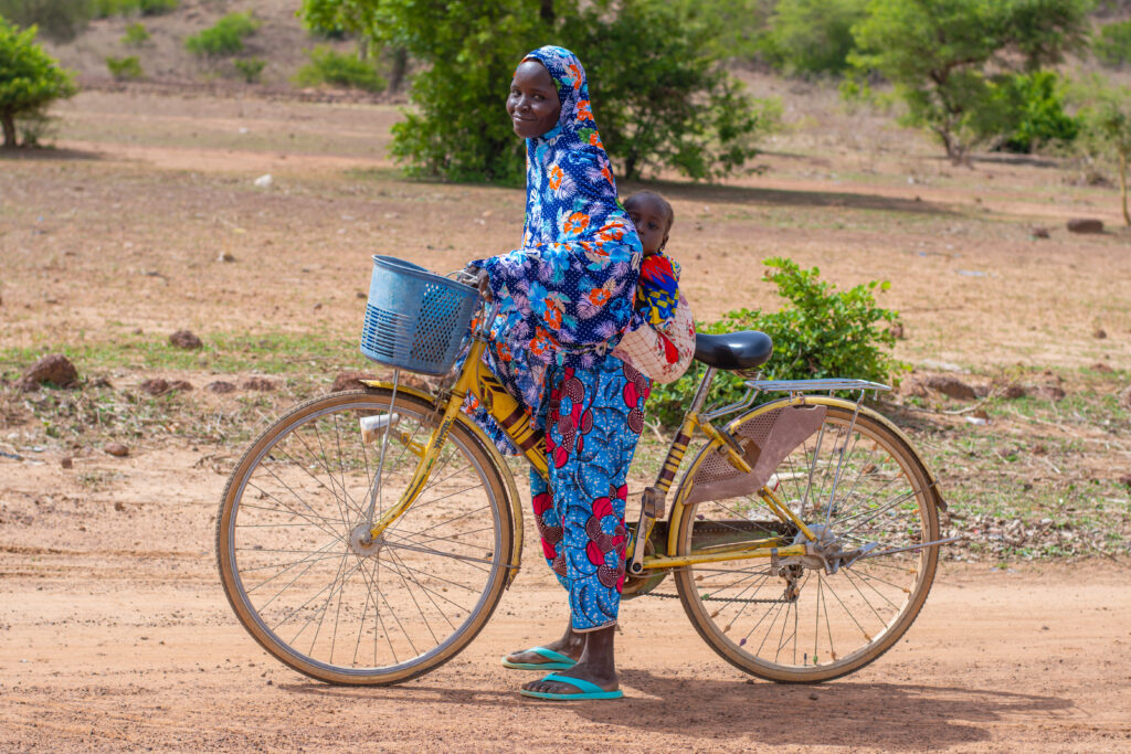 A woman in Burkina Faso with her baby on her back rides her bike.