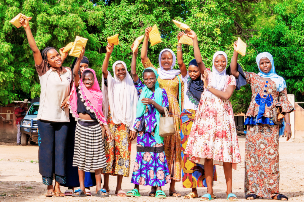 Group of young women and girls jump with joy while holding menstrual health and hygiene products they have helped create.