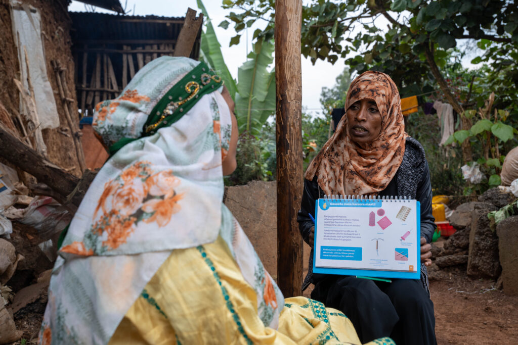 Two women sit next to each other, one holding a board with different contraceptive methods available for family planning.