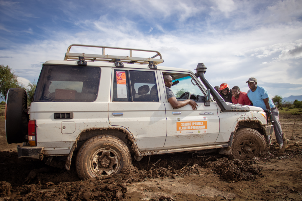 Photo of an SUV stuck in deep mud being pushed to safety by a team of EngenderHealth staff members